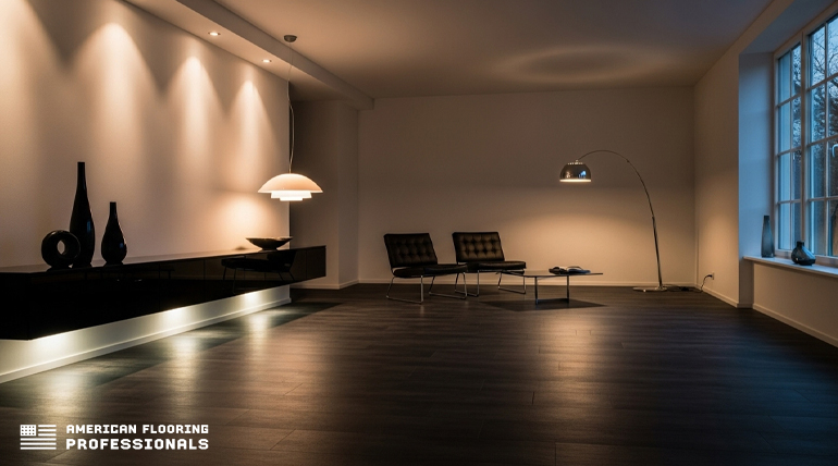 A dimly lit, spacious room with dark vinyl flooring. Recessed ceiling lights illuminate a white wall with accent lighting, highlighting three dark decorative vases and a bowl on a long, sleek black floating console with an illuminated strip underneath. A modern white pendant light hangs from the ceiling. In the middle distance, two black lounge chairs with a small table sit under the soft glow of a tall, curved floor lamp. A large window on the right reveals tree branches outside, with a dark vase and another decorative object on the windowsill. The bottom left corner features the "AMERICAN FLOORING PROFESSIONALS" logo.