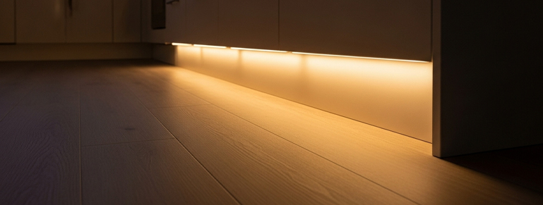 A low-angle close-up shot of warm LED strip lighting illuminating the kick plate area beneath white kitchen cabinets, casting a soft glow onto a light wood-grain vinyl floor.